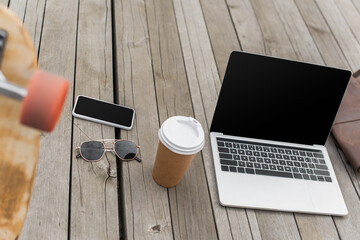 gadgets with blank screen, sunglasses and paper cup on wooden table near blurred longboard.