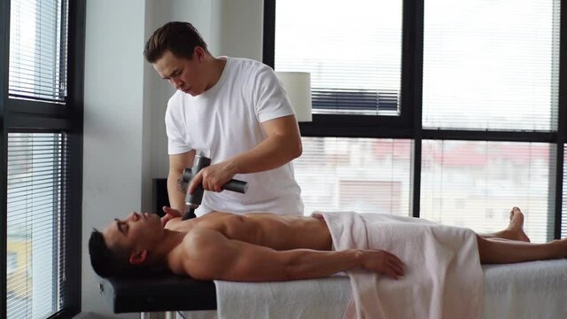 Side view of professional male masseur massaging chest muscles of muscular athlete with massage percussion device on background of window. Sportsman receiving electric lift massage in medic clinic.