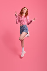 Full length body size photo o excited redhead woman jumping showing v-sign isolated on vivid pink color background, studio portrait. Young female in pink shirt and denim jeans looking happy