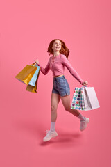 Full-length portrait of cute female with packages jumping high up celebrating weekend vacation, discounts in shop, black friday, wearing casual pink shirt and denim shorts, isolated on pink