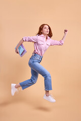 Fototapeta na wymiar Emotional redhead female student rushing forwards with book in hands, hurry to university, wearing casual outfit, running, 1 September exam, isolated in studio, full-length portrait