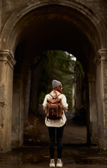 rear view on traveler woman with backpack stand in historical ancient arch, exploring interesting old sites and attractions, historical places, at autumn or spring season. view from back. full-length