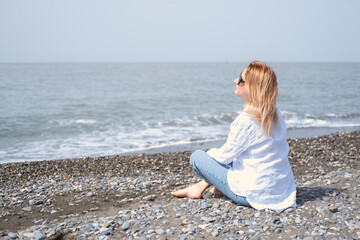 Fototapeta na wymiar Attractive young woman in white shirt and blue jeans with sunglasses sitting at beach