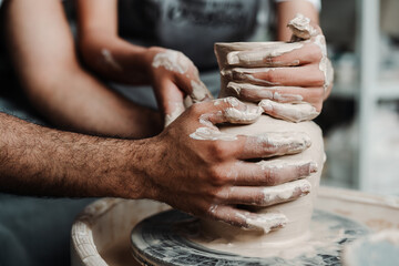 Photo from the side. Hands of a guy and a girl make a vase of clay on a potter's wheel