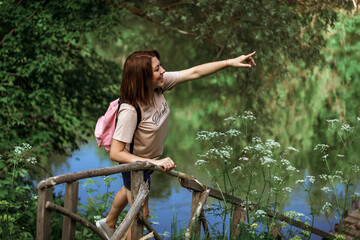 A happy young woman with short hair and a pink backpack stands on a small wooden bridge near the river in the forest, smiling and pointing into the distance. Selective Focus