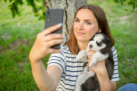 Young beautiful woman in a t-shirt and jeans with a husky puppy takes a photo or selfie on the phone while sitting in a park near a tree