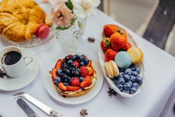 Piece of breakfast table outside with dessert, silver cutlery, coffee, croissant, macarons, blueberries, strawberries, flowers and anis stars