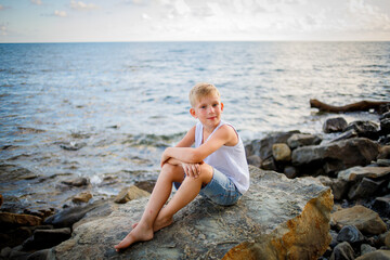 A boy in a white T-shirt and denim shorts sits on a rock near the seashore