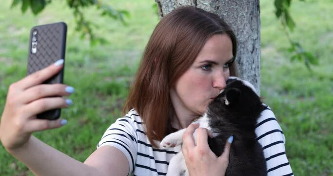 Young beautiful woman in a T-shirt and jeans with a husky puppy makes a photo or selfie, talks on the phone or shoots a video for social networks while sitting in a park near a tree
