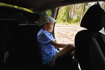 Focused boy sitting on a rear passenger seat in the opened door in the car and looking at the forest