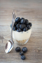 Healthy breakfast or dessert concept. Natural Greek yogurt with fresh blueberries and muesli in a transparent glass with a spoon on a wooden background. Menu for fitness, sport or healthy lifestyle.