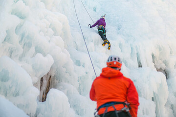Man standing and controlling a safety top rope while female with ice climbing equipment, climbing...