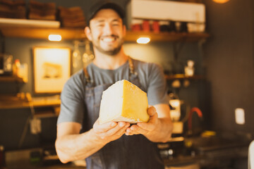Barista offering big nice piece of cheese with both hands in Brazilian organic artisanal bakery. Small business concept. Shop shelf out of focus on the background.