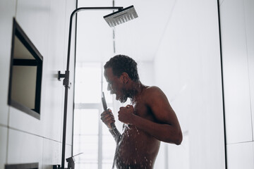 Positive young African-American man sings taking shower with hot water jets in contemporary unit in...