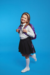 Adorable schoolgirl wearing uniform and backpack, smiling against blue wall