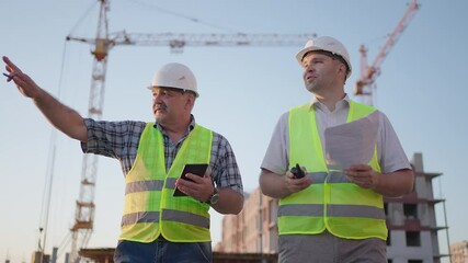Two engineers discussing project on a construction site, a worker wearing a helmet during the sunset