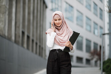Muslim business woman in formal clothes and hijab standing on street and talking on mobile. Confident female entrepreneur with clipboard in hands having working conversation on smartphone.