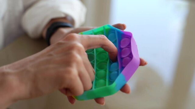 Close-up hands of unrecognizable young woman playing with multicolored pop-it fidget toy sitting at table. Closeup top view of female pushing colorful iridescent soft silicone bubbles sitting at desk.