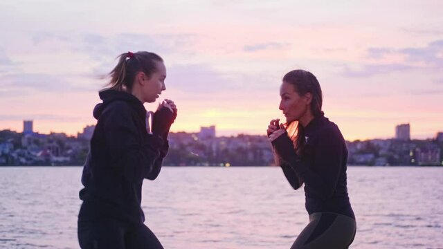 Two young women fighters practice side kicks and inclines against the background of a river at sunset. Sports, boxing