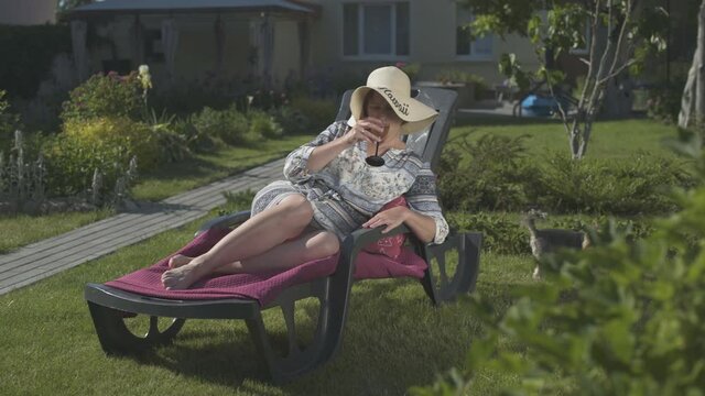 A woman in a straw hat lies on a sun lounger in the garden of her backyard, drinks wine and interacts with the Yorkshire Terrier.