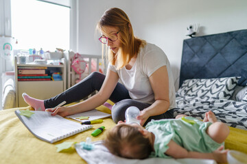 Front view of millennial caucasian woman mother sitting on the bed at home study while taking care...