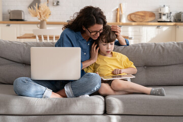 Happy mom embrace small kid playing games on tablet during break from remote work on laptop computer sit on sofa together. Cheerful young working mother bonding with preschool son on covid-19 lockdown