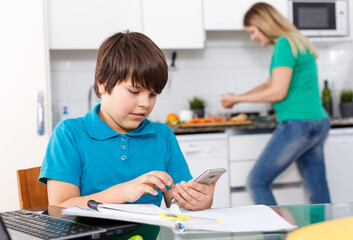 Positive schoolboy doing homework using laptop at kitchen interior, woman cooking at background