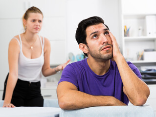 Upset young man sitting at home table with disgruntled woman behind