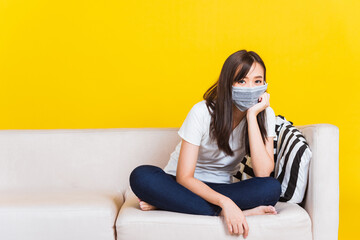 Portrait Asian of beautiful young woman sitting on sofa wearing medical face mask protective during Coronavirus studio shot isolated on yellow background