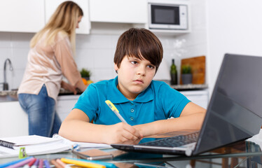 Portrait of boy studying at kitchen table and doing homework while his mother cooking