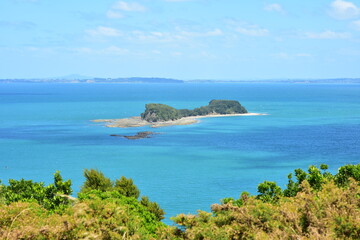 View of Saddle Island in blue waters of Hauraki Gulf with reefs along shoreline exposed during low tide.