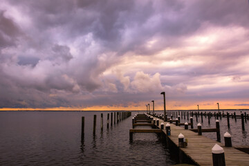 Fototapeta premium Storm clouds in the distance getting lit up with stunning sunset colors over a dock. Island Beach State Park New Jersey