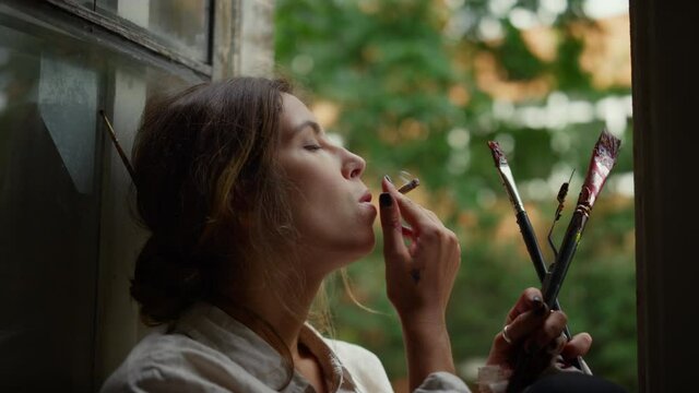 Creative woman holding cigarette indoors. Female painter smoking on windowsill.