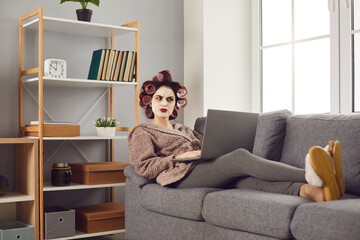 Funny angry woman in hair curlers sitting on sofa, using laptop computer, posting negative customer...