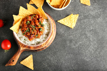 Bowl with tasty chili con carne, rice, nachos and tomatoes on dark background