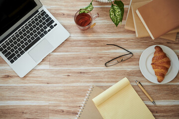 Desk of student with opened laptop, books, planner, cup of tea and croissant