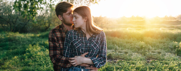 bearded guy kisses a girl standing in the spring forest or in the park at sunset. Loving couple husband and wife hugging. Man and woman romantic date. Vacation in nature. Banner.