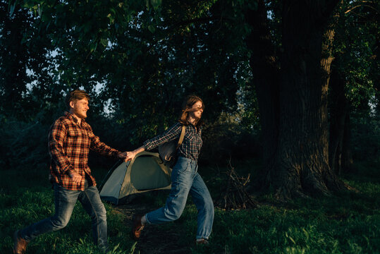 happy guy and girl travelers run near the tent in the forest at sunset in the evening. A couple of young lovers tourists in the autumn or spring forest at the campsite. People outdoors.