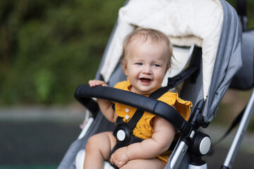 Sweet caucasian baby girl ten months old sitting in stroller outdoors. Little child in pram. Infant kid sits in pushchair. Summer walks with kids. Family leisure with little child.
