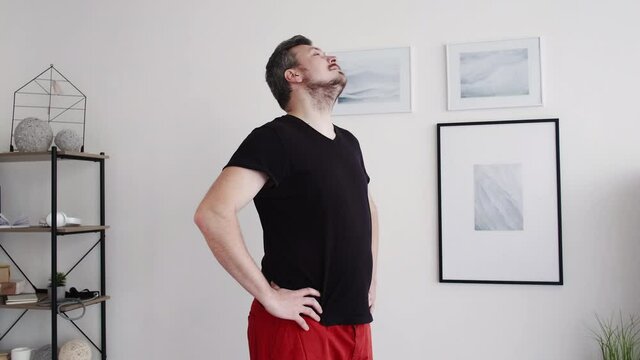Healthy sport. Chubby man. Home fitness. Keep fit. Middle-aged happy guy in homewear sportive t-shirt shorts doing warming workout neck hands in light room interior.