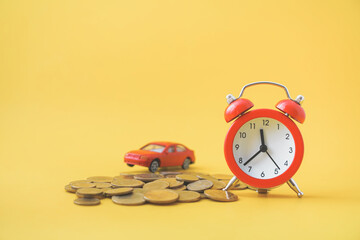red analog alarm clock , blurred miniature car on pile of coins on yellow background, business and...