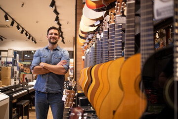 Portrait of young talented musician standing in music shop by large collection of guitar...
