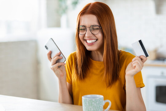 Young cheerful caucasian girl doing online shopping at home using mobile phone and credit card, redhead beautiful excited girl feels euphoria from a long-awaited purchase the Internet, smiling