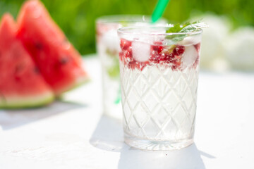 drink with pomegranate berries, green currant leaves and ice cubes, on a white wooden background, watermelon slices in the background, sunny day.