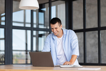 Fototapeta na wymiar Successful young caucasian businessman using laptop for work standing in the modern office, smiling