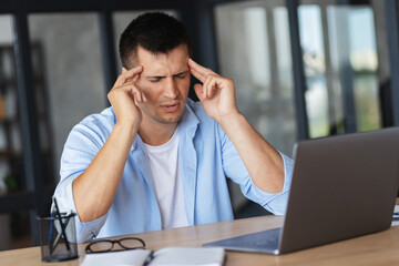 Tired caucasian businessman having headache or migraine sitting at the office desk. Young man freelancer suffers from headaches after working at the computer for a long time