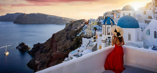 A tourist woman in a red dress and hat sits on a balcony in the village of Oia, Santorini, Greece,...