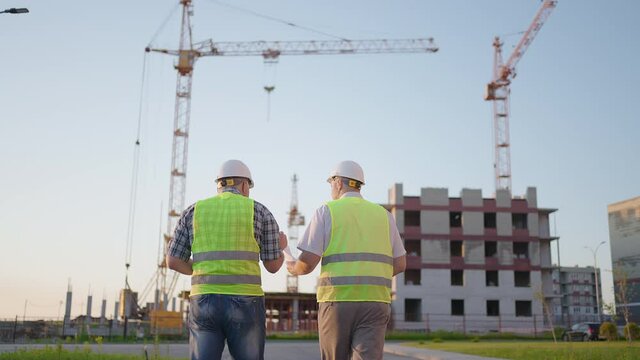 Waist-up of two middle-aged male builders wearing safety clothing, standing at construction site, man using walkie-talkie, his colleague holding paper with project plan