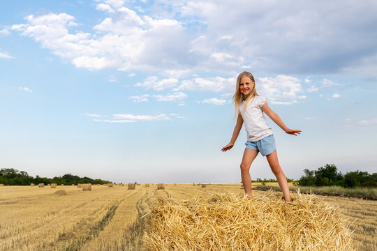 Portrait of cute little blond beautiful adorable cheerful caucasian kid girl enjoy sitting on hay stack or bale on harvested wheat field warm summer evening. Scenic rural country landscape background