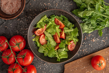 There is a bowl of light summer vegetable salad of tomatoes and greens and fresh ingredients for its cooking on a black background. Top view